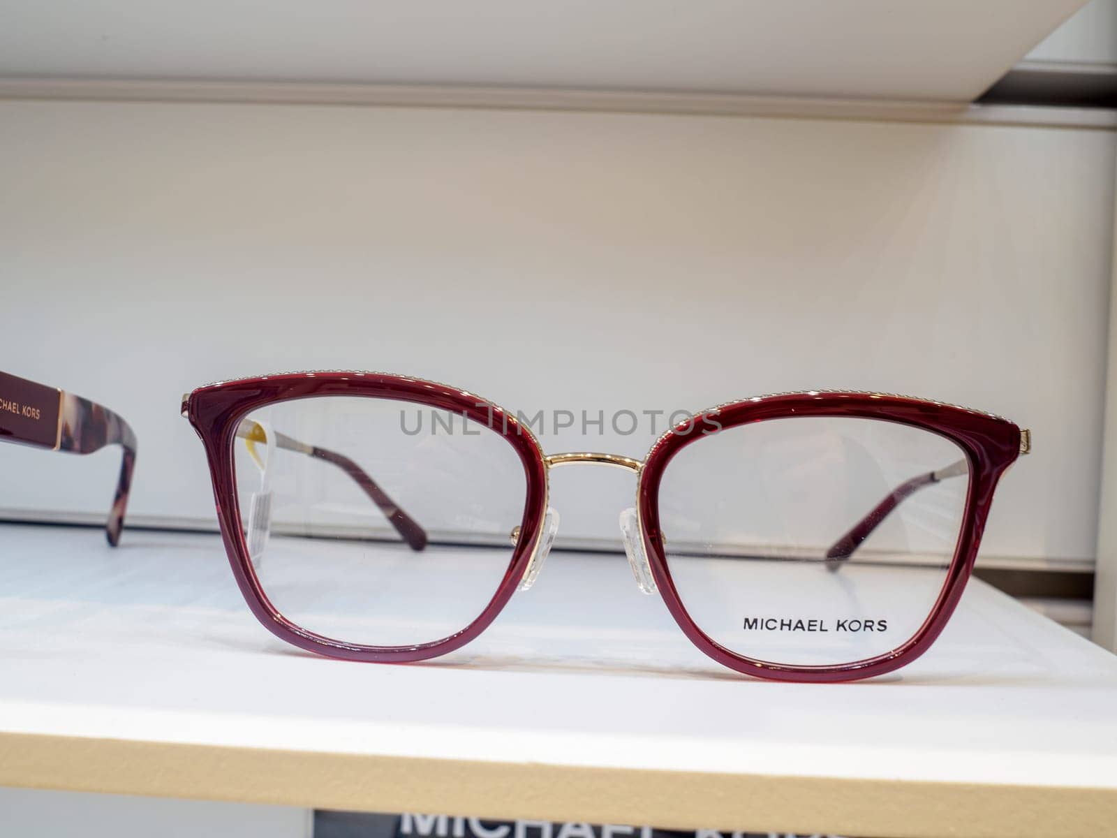 Stylish michael kors eyeglasses with dark red frames are displayed on a white shelf in an eyewear store, ready for customers to try on