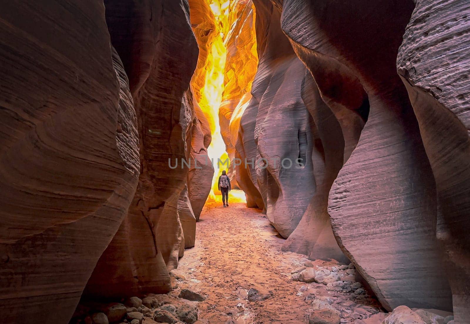 A female hiker walking through the narrow Buckskin Gulch slot canyon on the Arizona, Utah border. She hikes toward the light at the end of the canyon with the tall canyon walls glowing orange - USA