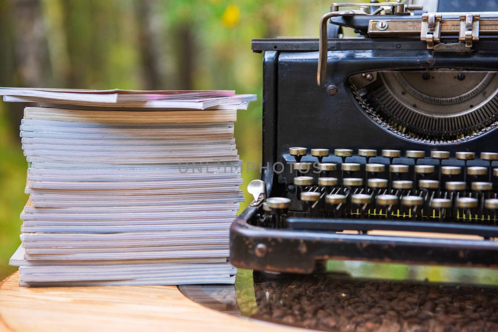 Close-up of an old-fashioned typewriter beside a tall stack of papers, set outdoors with a blurred natural background.