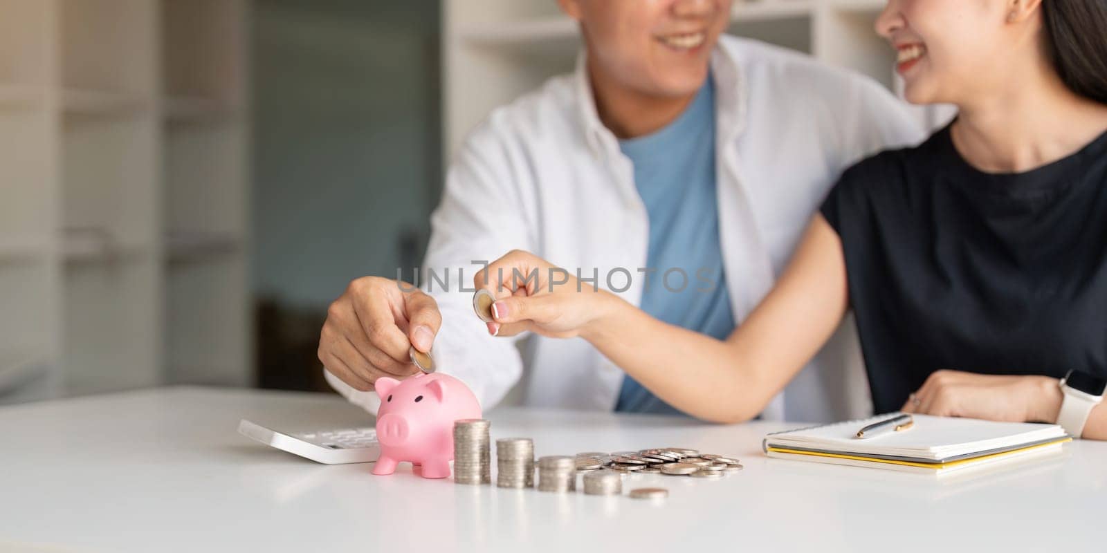 Family savings. Happy couple asian putting coin in piggybank while sitting together on desk at home, loving spouse planning budget and saving money for future by nateemee