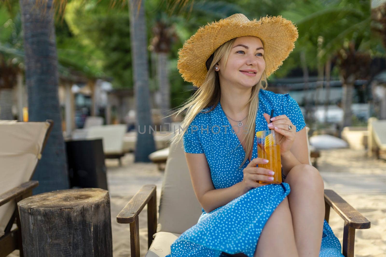 Fashion young woman drinking cocktail in beach bar. Cheerful attractive blonde woman in a dress smiles and drinks a cocktail outdoors. Happy woman enjoying summer time at the beach.