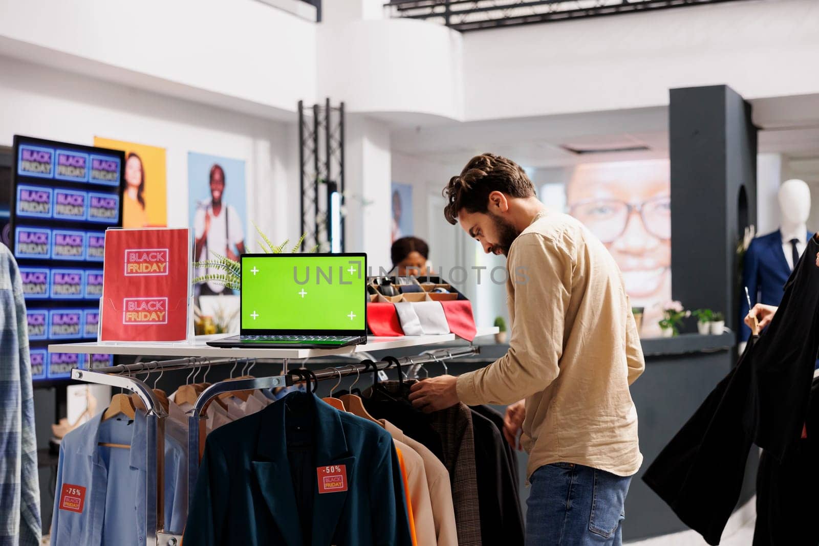 Seasonal discounts. Young man shopping on Black Friday in fashion boutique, looking for discounts during holiday season sales. Laptop with chroma key green screen standing on clothing rack