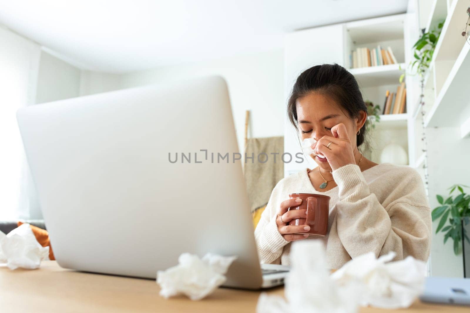 Young Chinese woman feeling sick working from home office. Asian female blowing nose with tissue holding cup of healing herbal tea. Wellness concept.