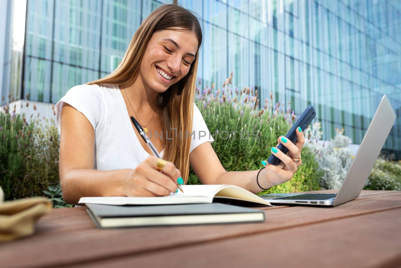 Young happy blonde female college student using phone and laptop to study, taking notes on notebook. University, college concept.