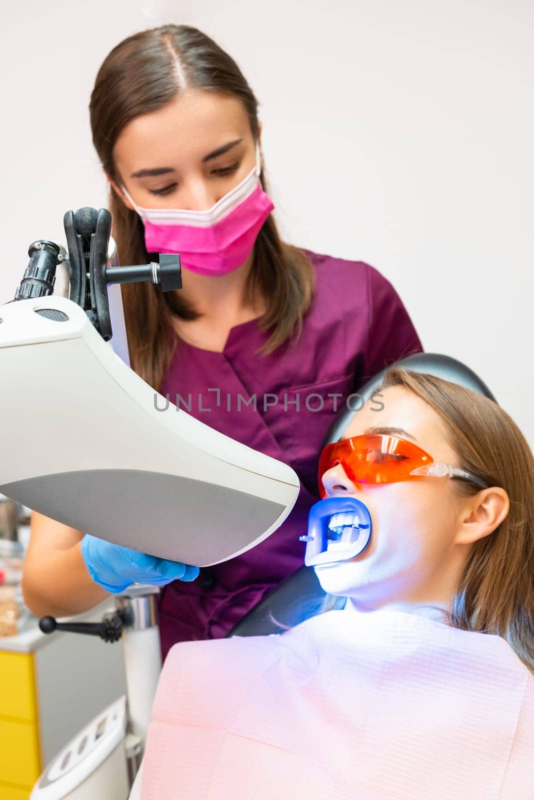 Young dentist in face mask performs whitening of teeth using UV lamp for woman at dentist clinic, vertical photo