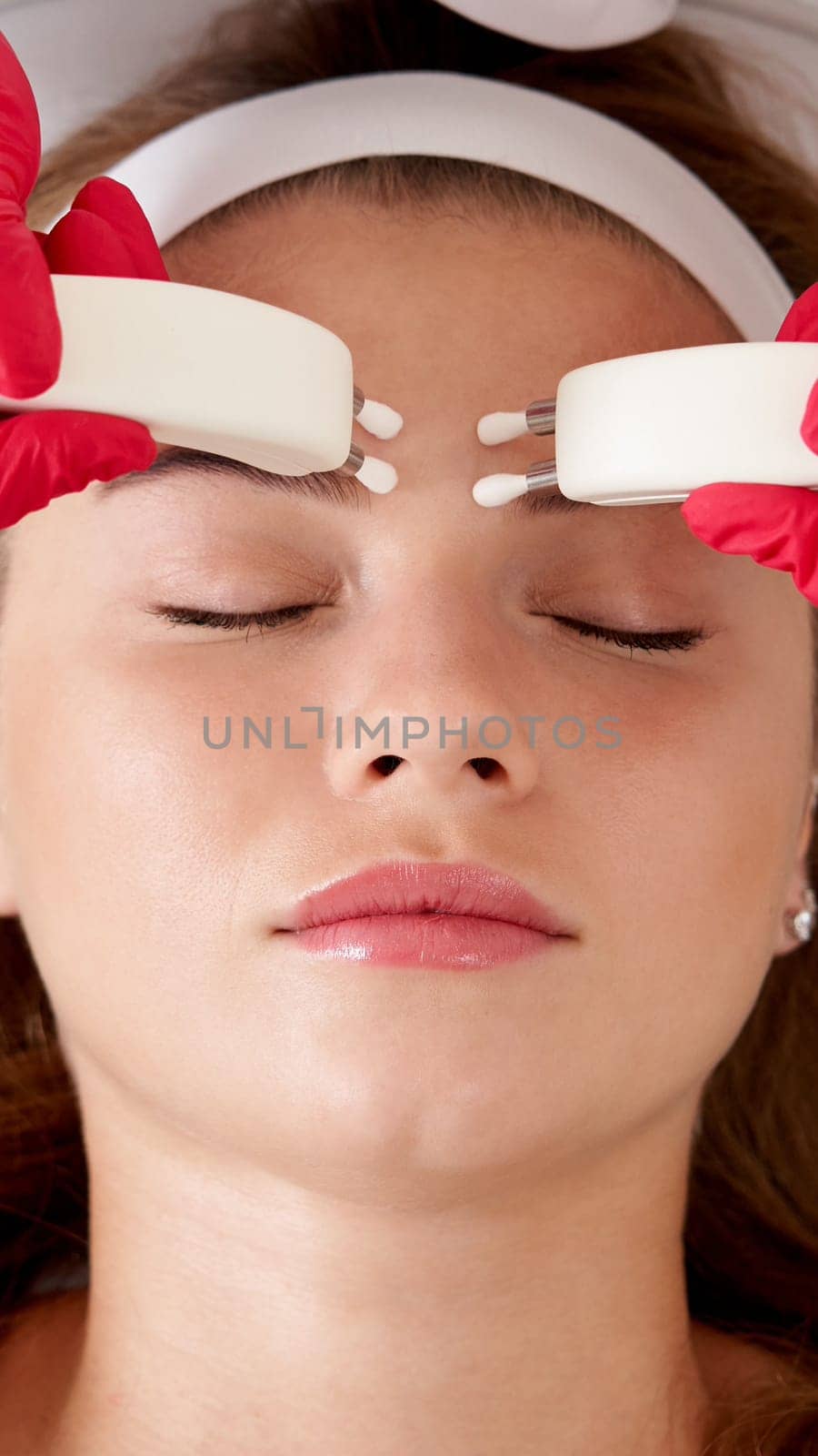 Beautiful Woman in Spa Clinic Receiving Stimulating Electric Facial Treatment From Therapist. Closeup Of Young Female Face During Microcurrent Therapy by Mariakray