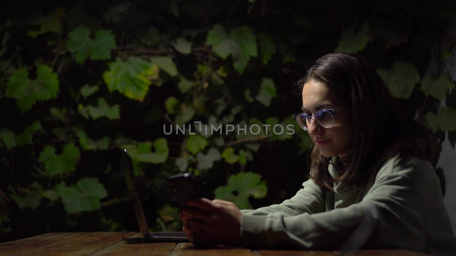 A young woman at a table in a gazebo is texting on a smartphone late at night. A girl in a hoodie and glasses with a phone in her hands against a background of green grapes. 4k