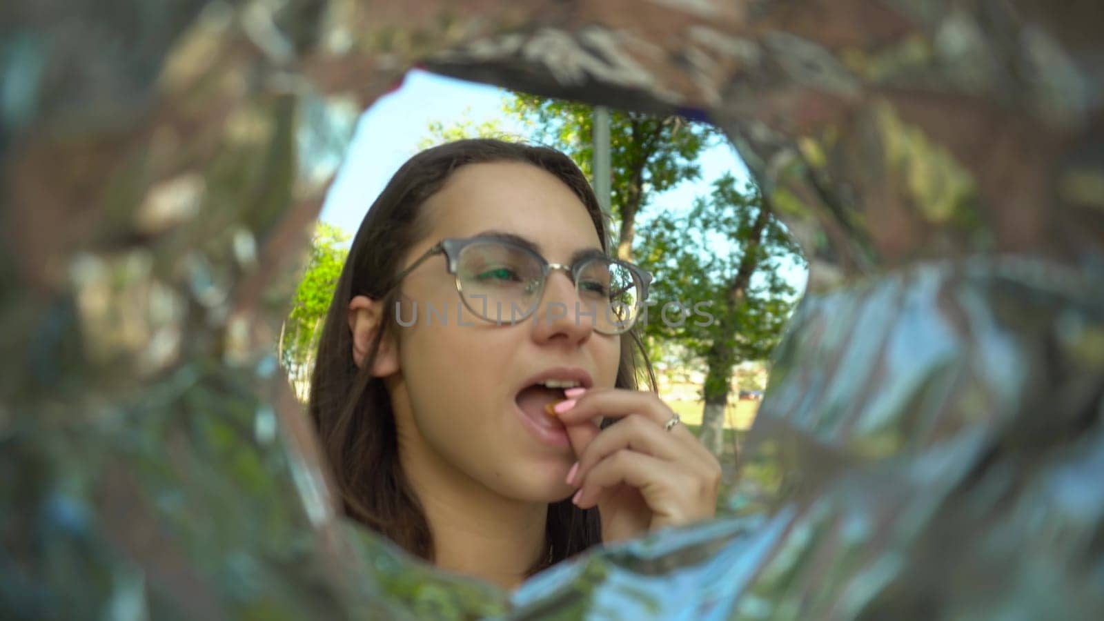 A young woman with glasses eats potato chips from a pack while walking in the park. View of a woman from a bag of chips. 4k