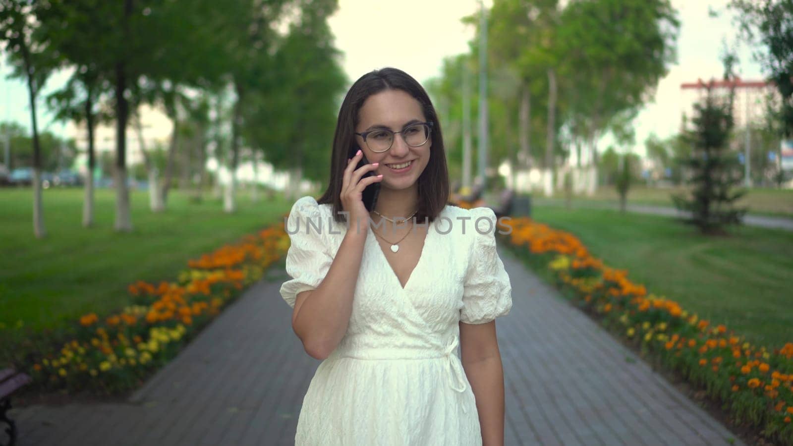 A young woman is walking in the park and talking on the phone. A girl in glasses and a white dress walks along a green alley. 4k