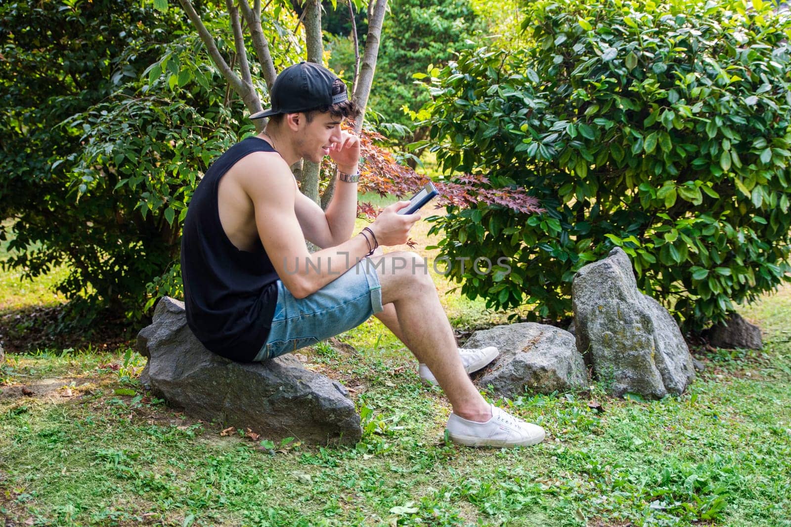 A man sitting on a rock in the grass, reading from an ebook reader. Photo of a handsome young man sitting on a rock in the grass with an e-book reader