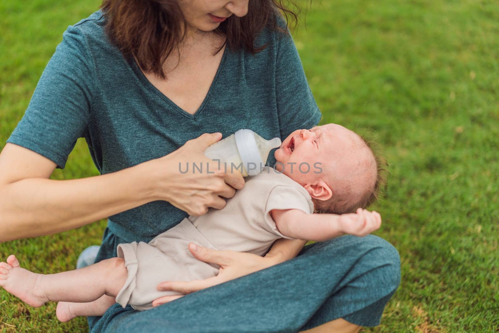 Mother holding and feeding baby from milk bottle in the park. Portrait of cute newborn baby being fed by her mother using bottle. Loving woman giving to drink milk to her boy.