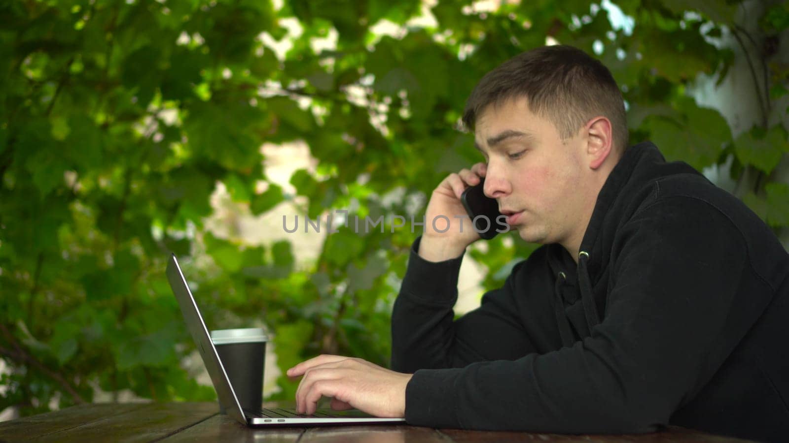 A young man at a table in a gazebo talks on the phone during the day. A man in a black hoodie with a laptop against a background of green grapes. 4k