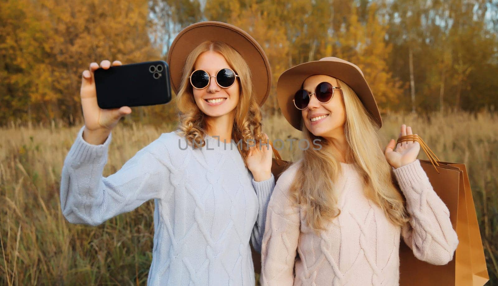 Happy two women friends taking selfie with phone in autumn park, girlfriends smiling holding shopping bags together outdoors in warm fall season