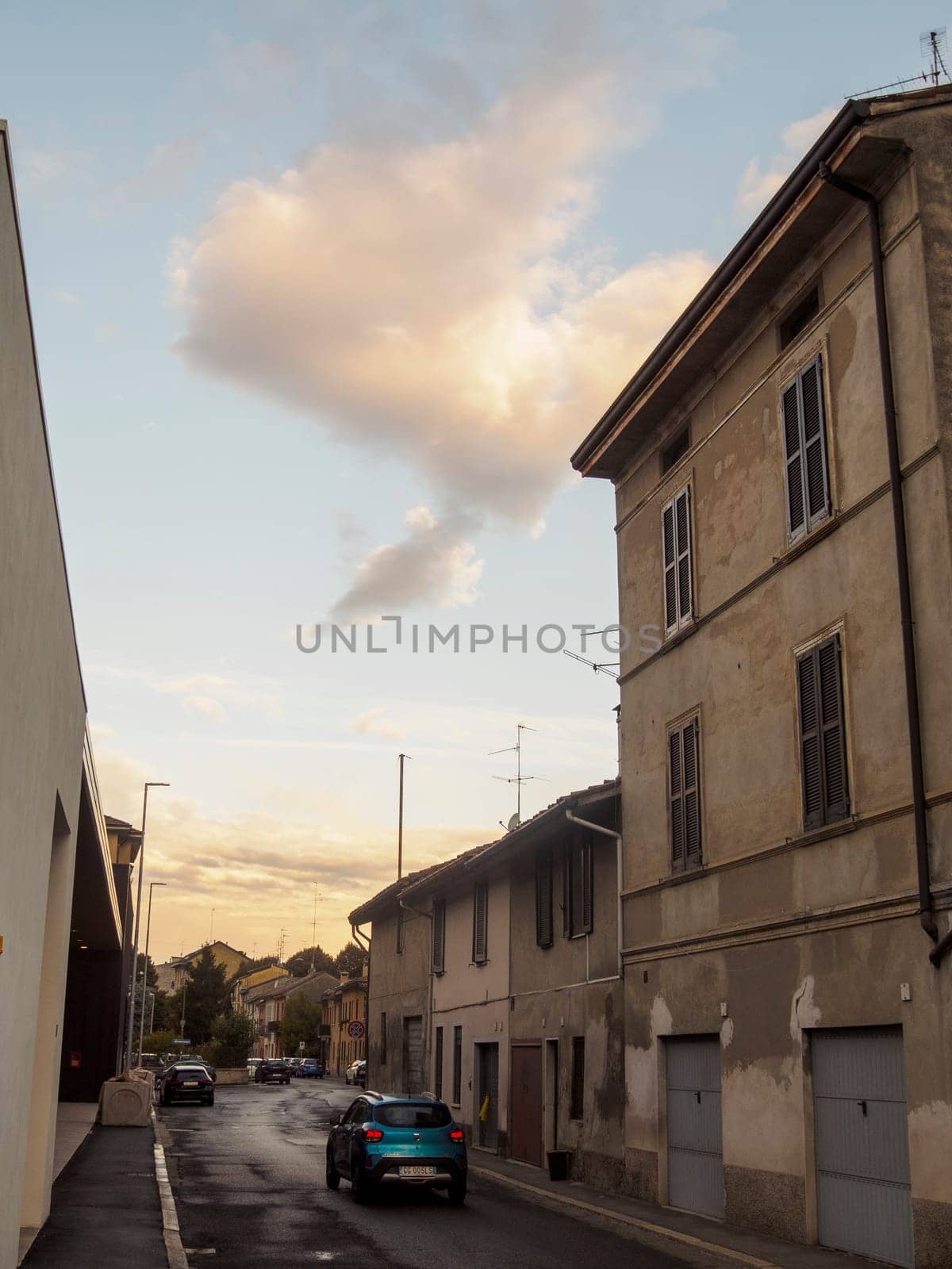 Cremona, Italy September 26th 2024 A blue car is driving down a narrow street in an italian village at sunset, with old buildings and a cloudy sky