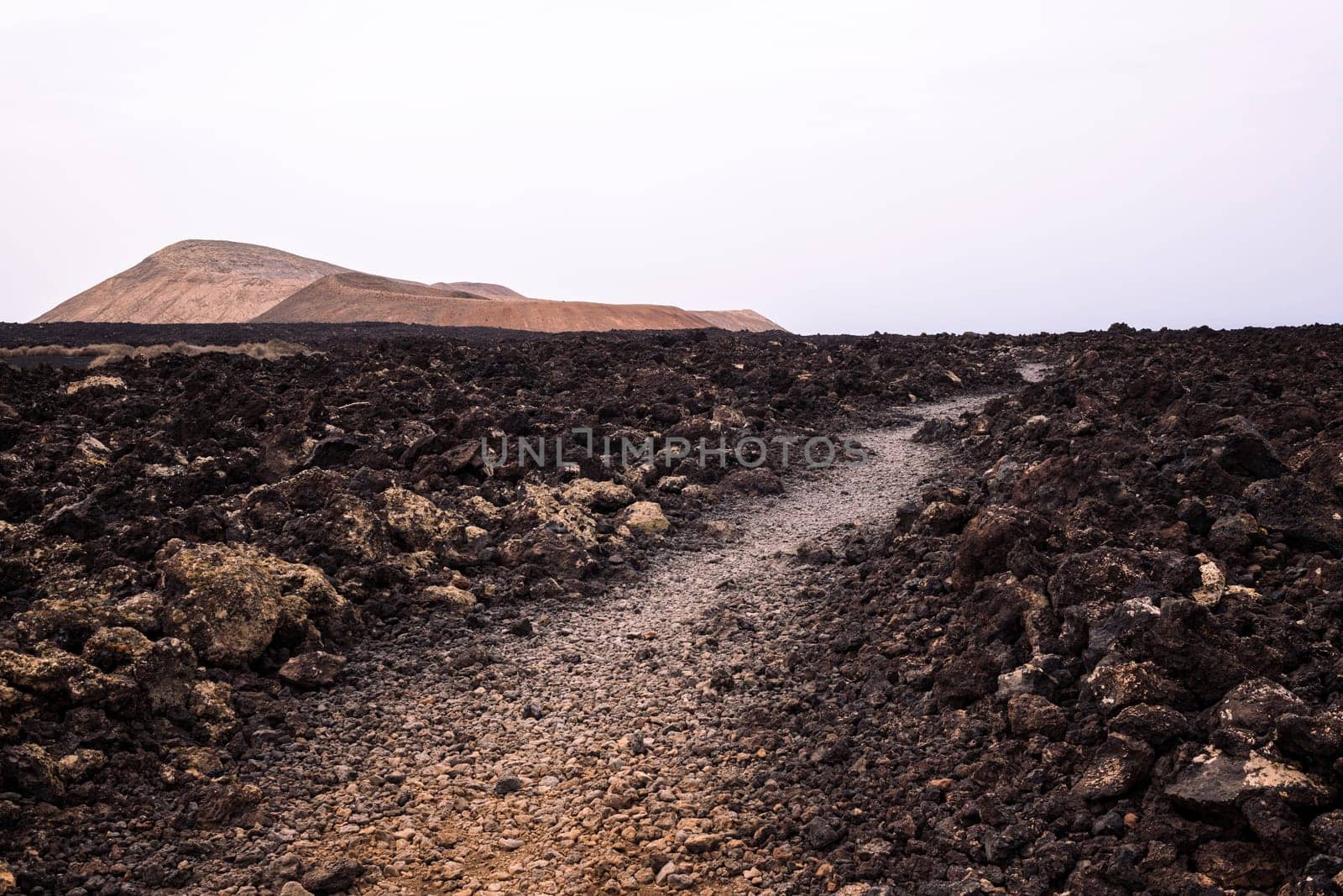 rough route with trail between dark lava stones against Caldera Blanca and Caldereta volcano in Natural Park of Lanzarote in Canary Islands, Spain, concept of volcanic landscape and wild nature