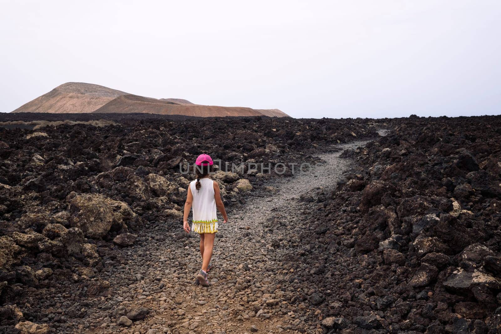 rear view of unrecognizable girl walking by rough trail between lava rocks and volcanoes in Natural Park of Lanzarote in Canary Islands, Spain, concept of active tourism and adventure travel