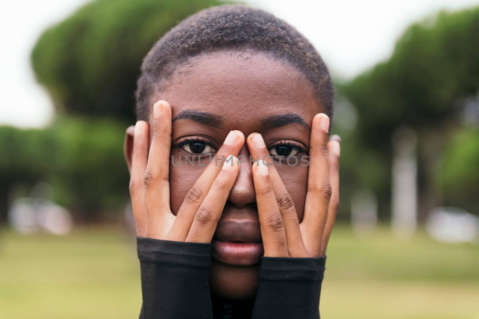 young tender african american female with short hair covering face with hands while looking at camera in a town park on summer day