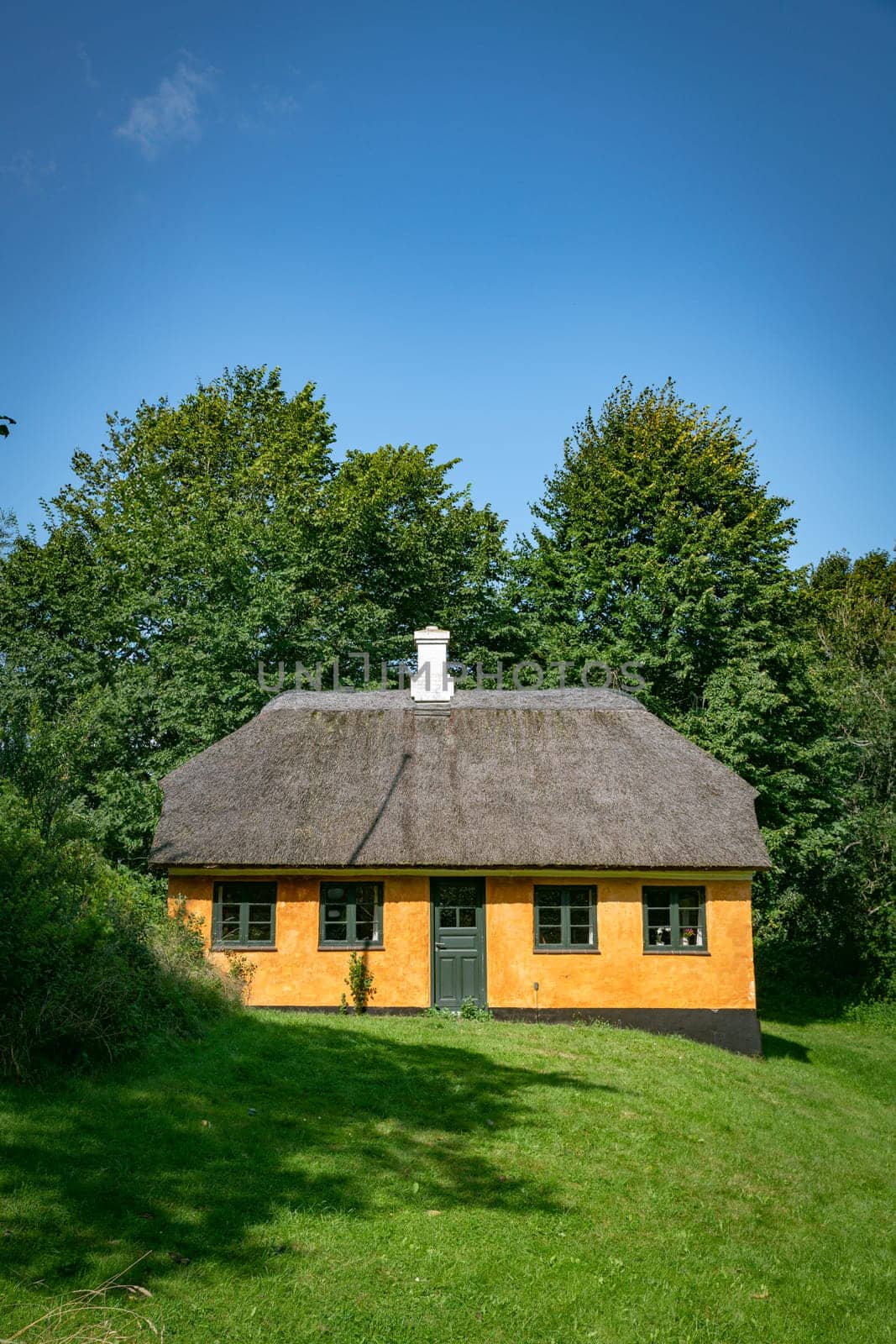 A picturesque orange cottage with a traditional thatched roof surrounded by lush greenery under a clear blue sky. Perfect depiction of rural tranquility and rustic charm.