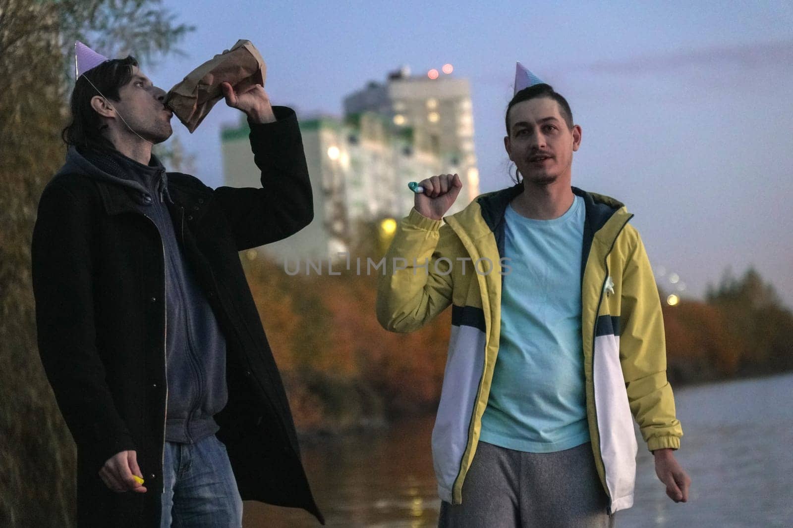 Two friends stroll along the water edge at twilight, wearing festive hats and sharing laughter.
