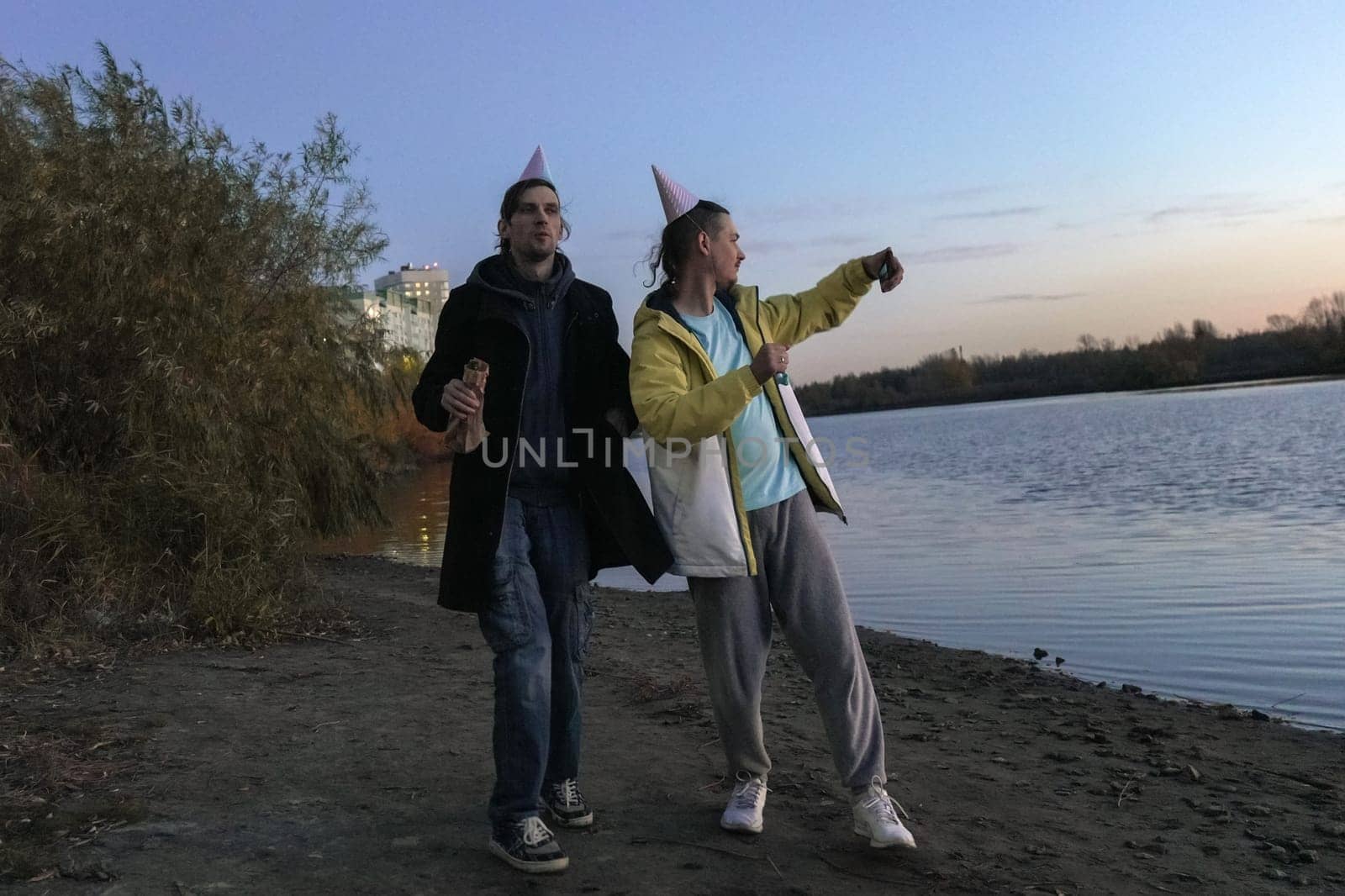 Celebrating friendship by the riverside at dusk with laughter, hats, and snacks during a joyful holiday gathering