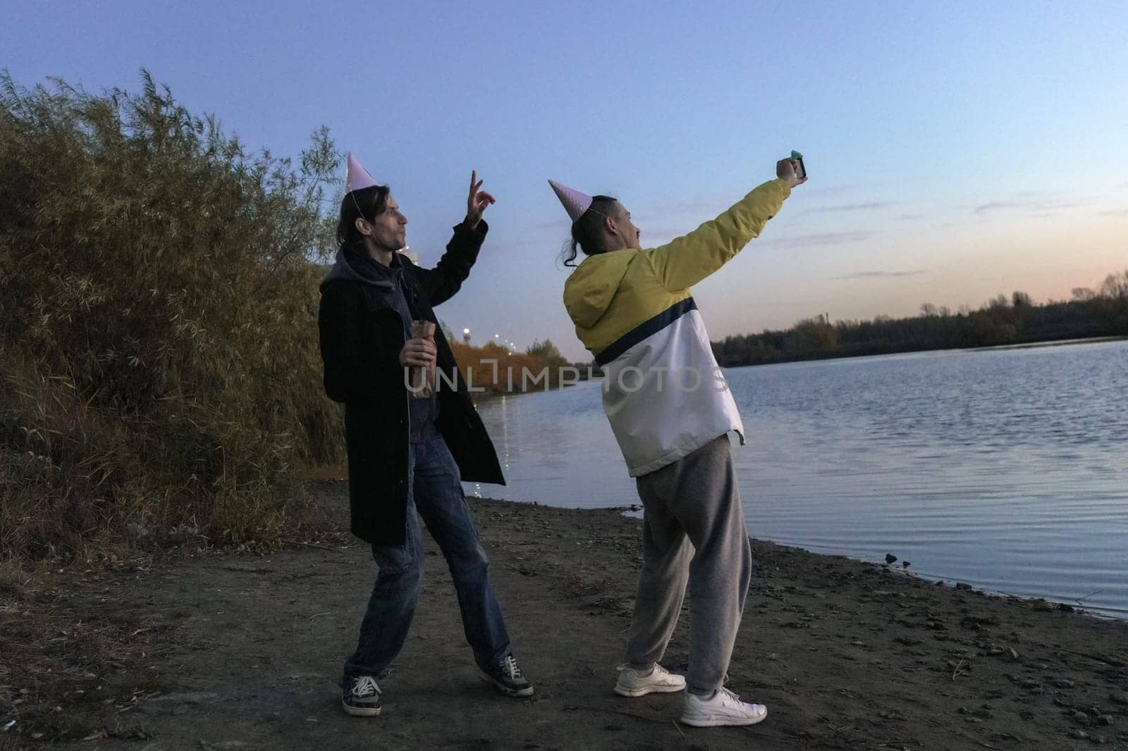 Two friends celebrating a holiday by the water at sunset, capturing joyful moments with festive hats and laughter in the air