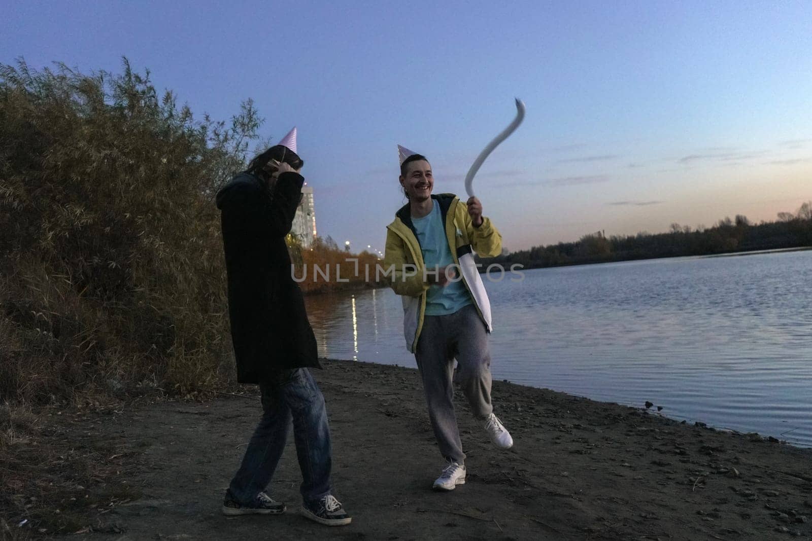 Two friends stroll along the water edge at twilight, wearing festive hats and sharing laughter.