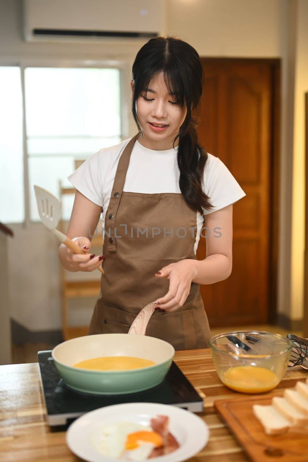 Young woman wearing a brown apron cooking scrambled eggs in a green frying pan.