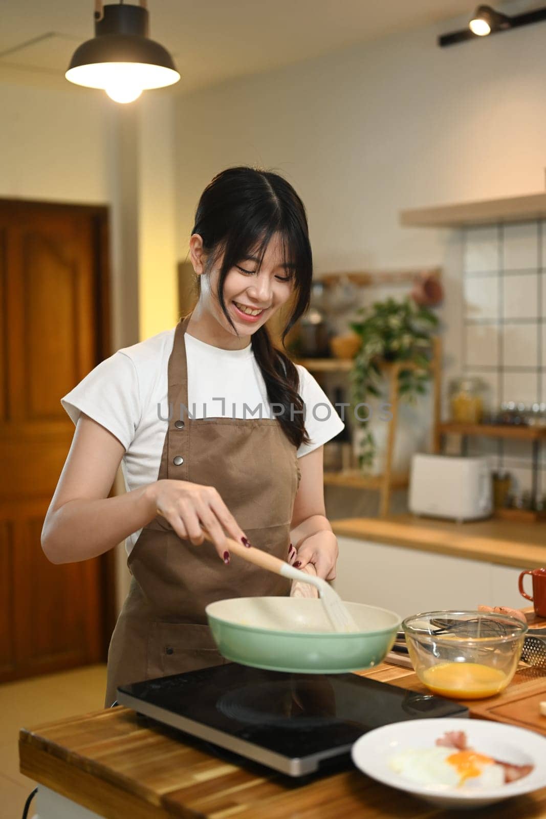 Cheerful young asian woman cooking scrambled eggs in frying pan on a stovetop.