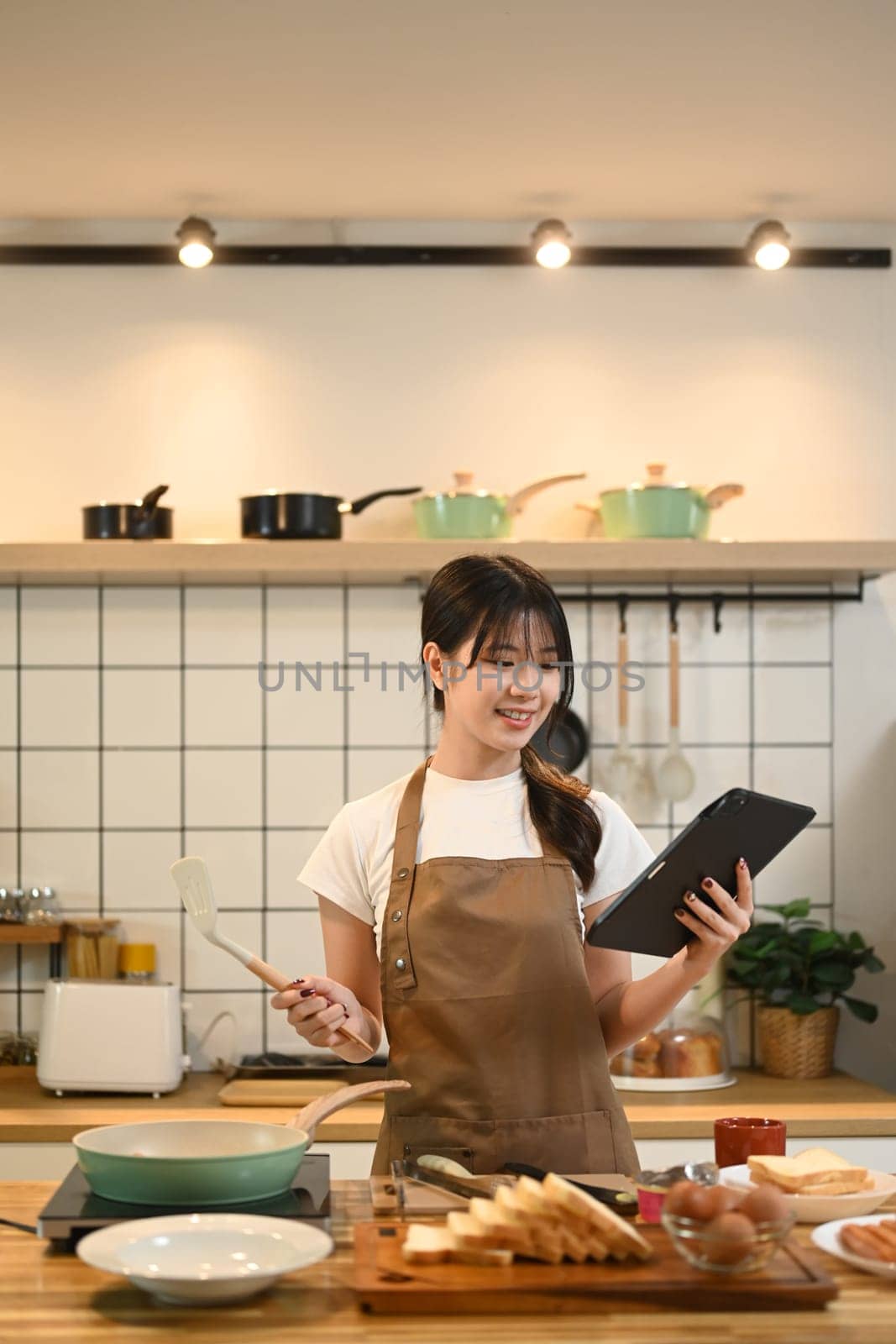 Smiling young woman standing in a kitchen, following a recipe or cooking tutorial online on digital tablet.