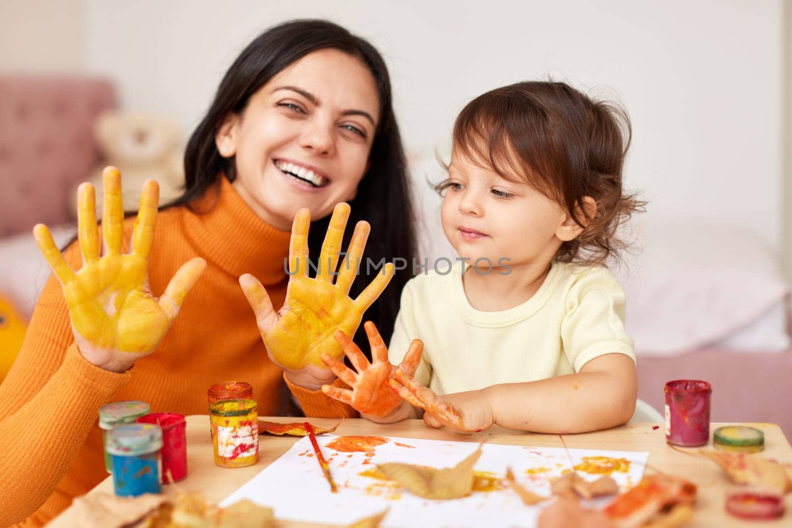 happy little child girl and mother painting hands and autumn leaves together. smiling mom and baby showing hands in yellow paint