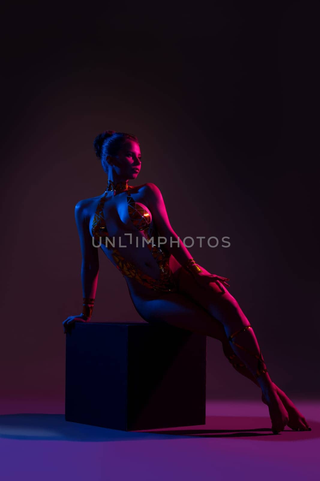 Beautiful nude girl with gold tape body art sitting on a cube in dark room