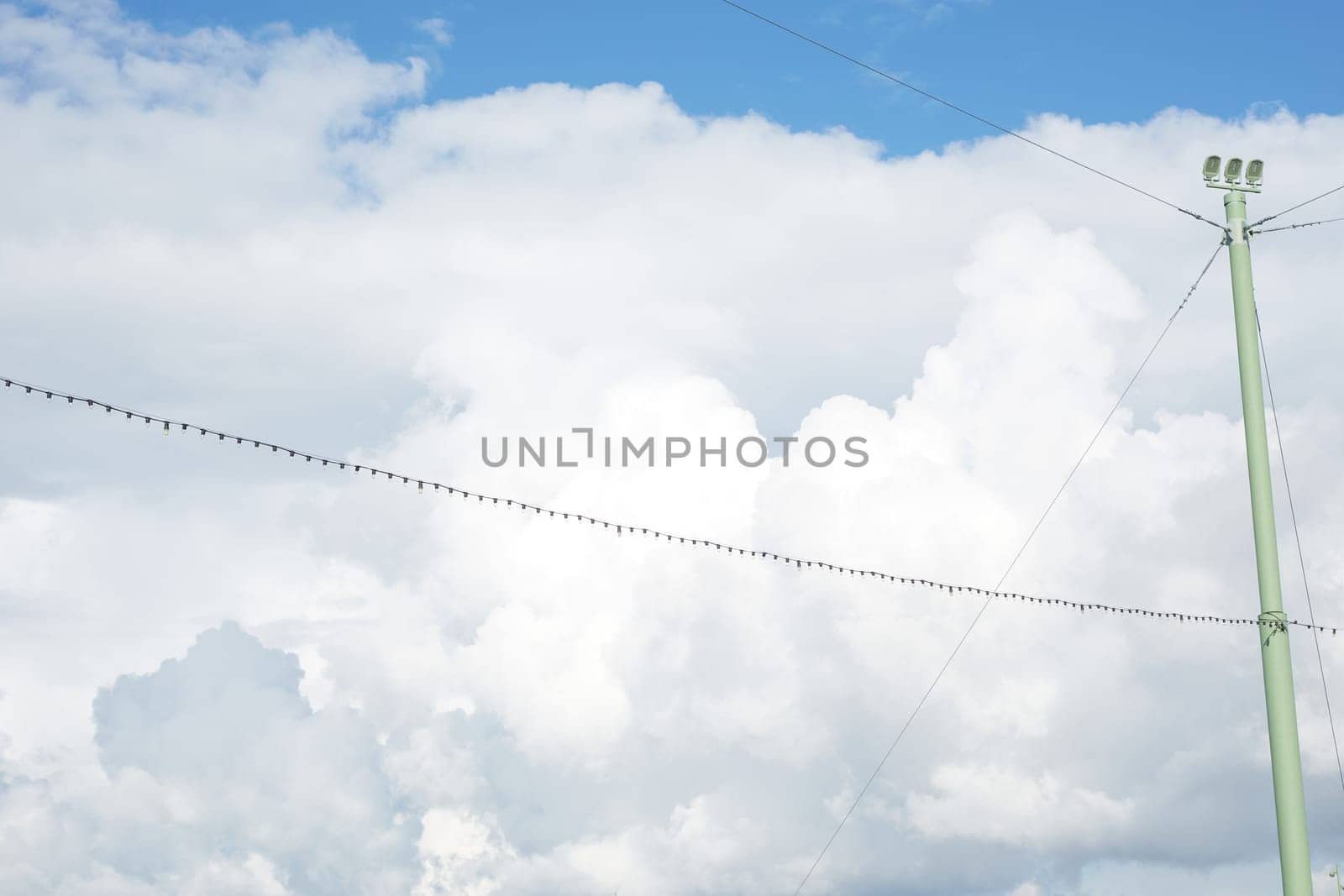 A calm blue sky with white clouds, a garland of electric bulbs and a pillar of street lighting.