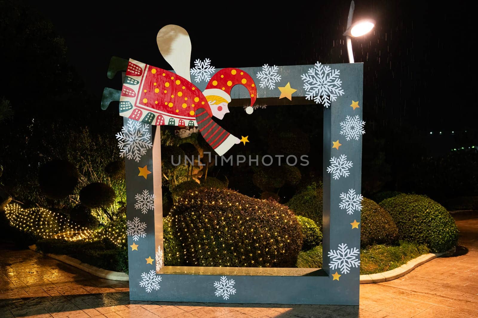 A whimsical Santa Claus figure is positioned above a large decorative frame in a beautifully lit garden. The vibrant colors and festive designs add a joyful atmosphere during the holiday season.
