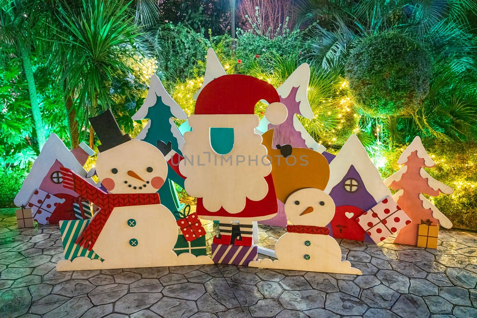 Festive decorations showcase Santa Claus with snowmen and colorful houses, surrounded by lush greenery and twinkling lights in a cheerful holiday garden.