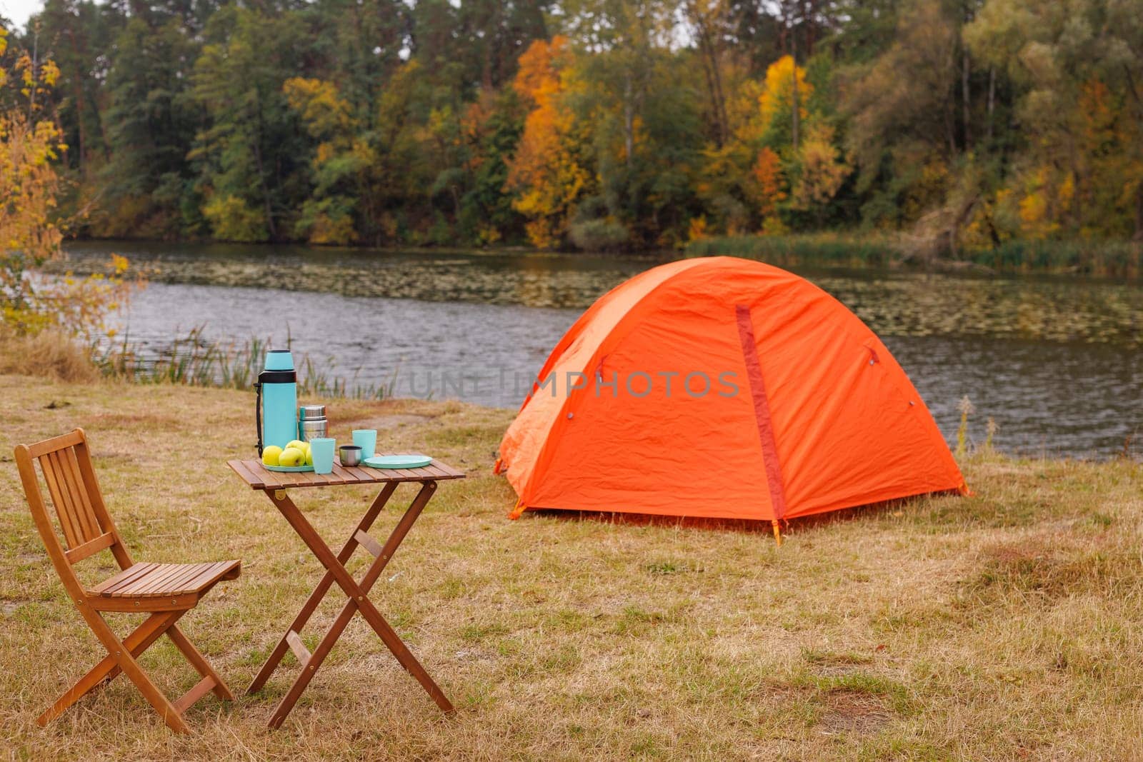 A vibrant orange tent set up by a serene riverbank, surrounded by autumn foliage. A wooden table with a thermos, cups, and apples is nearby, creating a cozy camping scene.