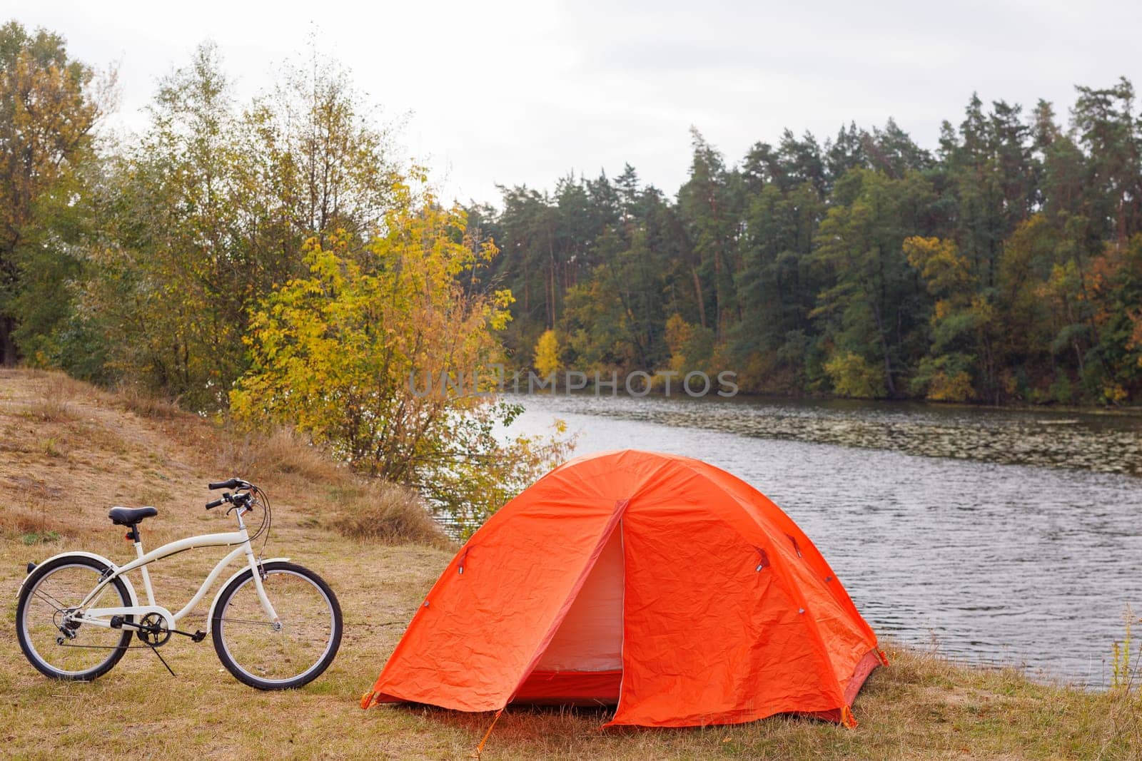 orange tent set up on grassy lake shore, bicycle in front of it, bicycle tourism, green tourism concept
