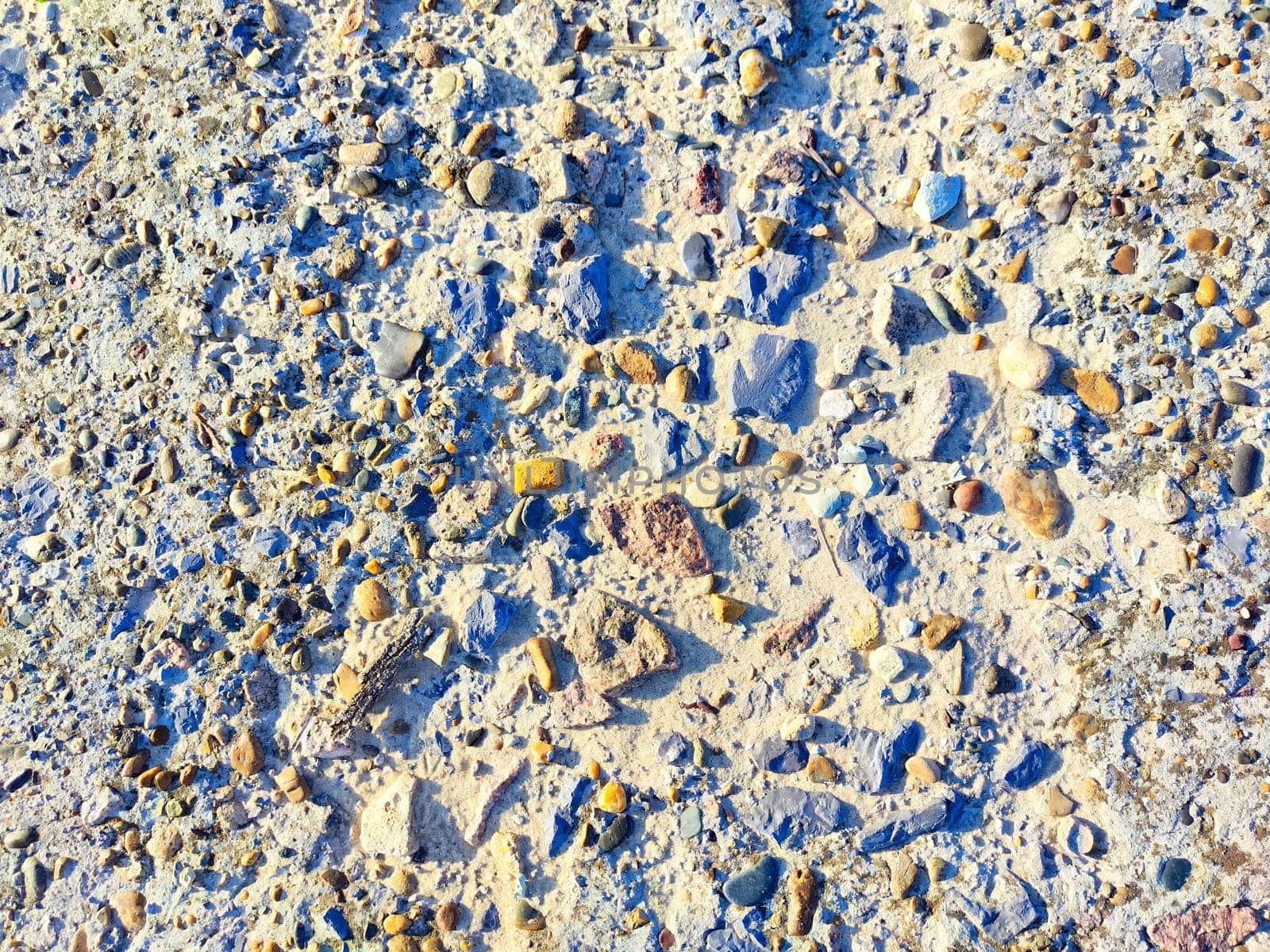 A variety of smooth stones in different colors are scattered on a sandy ground, illuminated by sunlight in an outdoor environment.