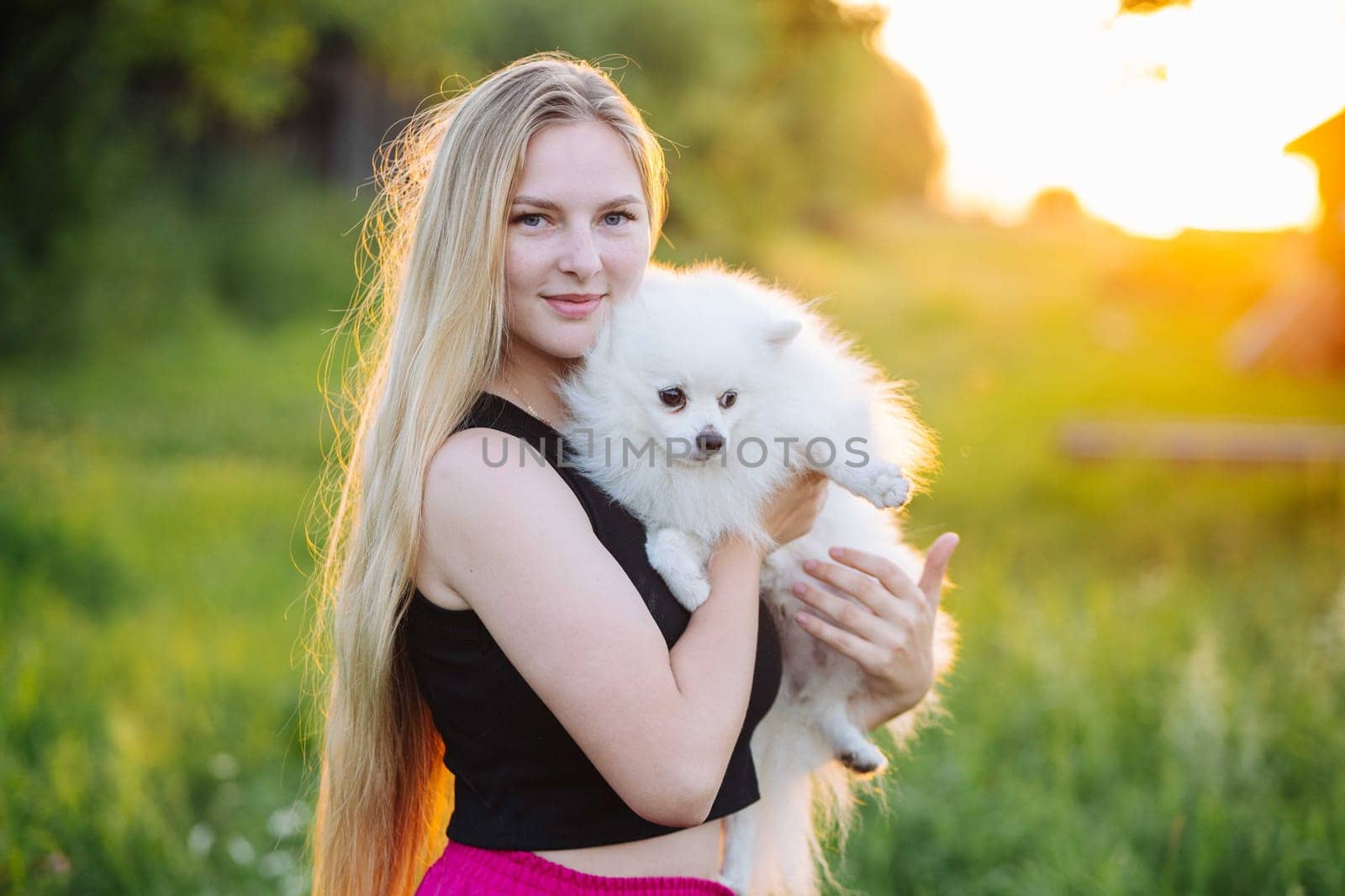 Blonde young woman is holding her white fluffy dog in the evening sun with a meadow in the background