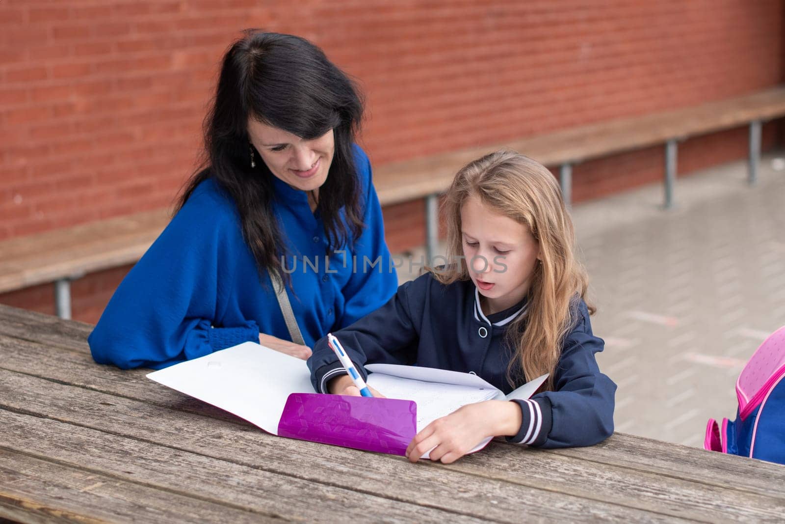 loving mother watching her daughter doing homework on her own at wooden table in children's playground, education concept, back to school, girl in beautiful blue uniform,