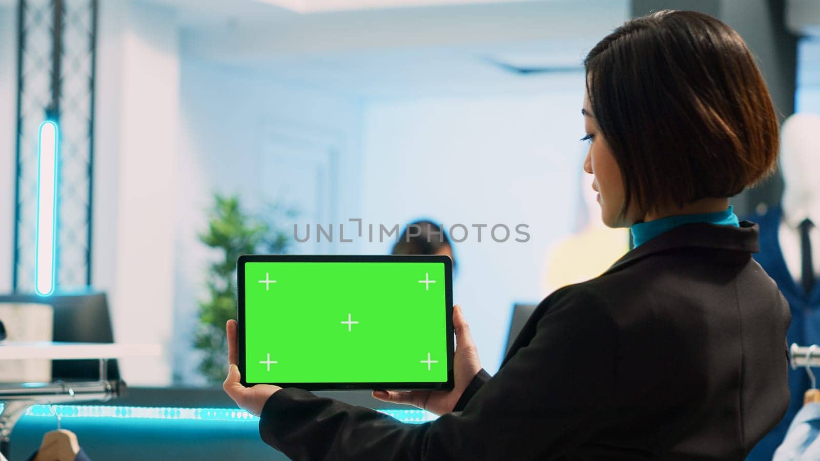 Female store assistant using tablet with greenscreen, working in clothing store. Young woman holding device with isolated display and chroma key mockup template, blank copyspace.