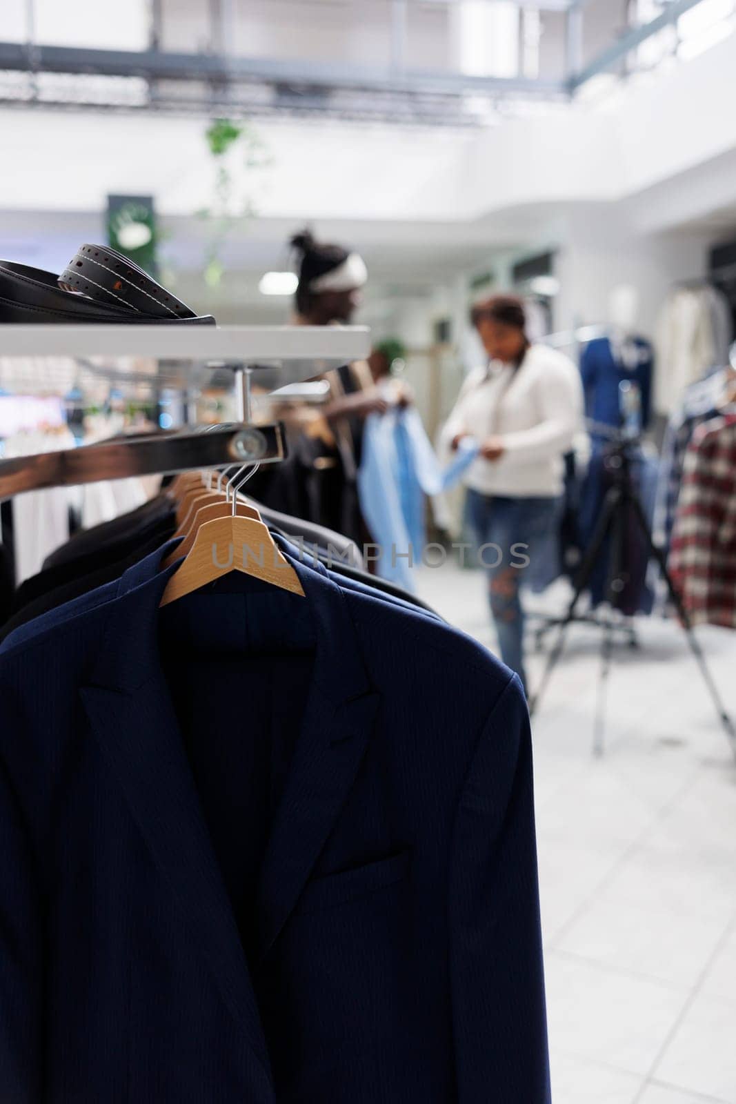 Formal male blazers hanging on rack in fashion boutique with blurred background. Male jackets latest arrival on hangers and accessories on shelf in department shopping mall close up selective focus