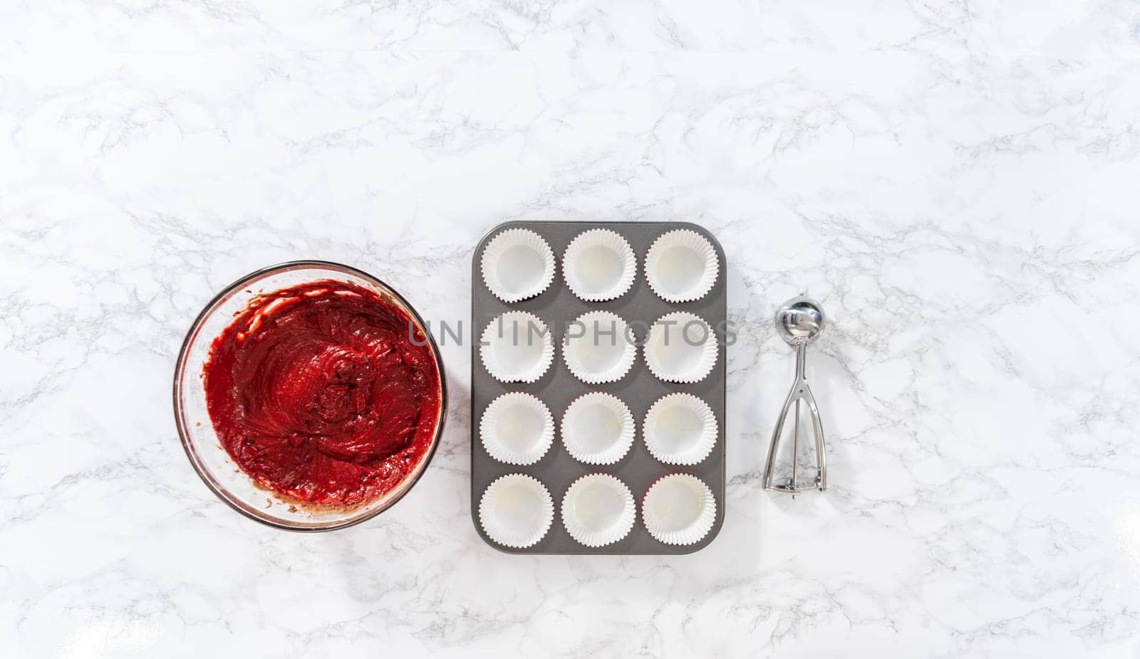 Flat lay. Scooping cupcake dough with dough scoop into cupcake pan lined with foil liners to bake red velvet cupcakes with white chocolate ganache frosting.