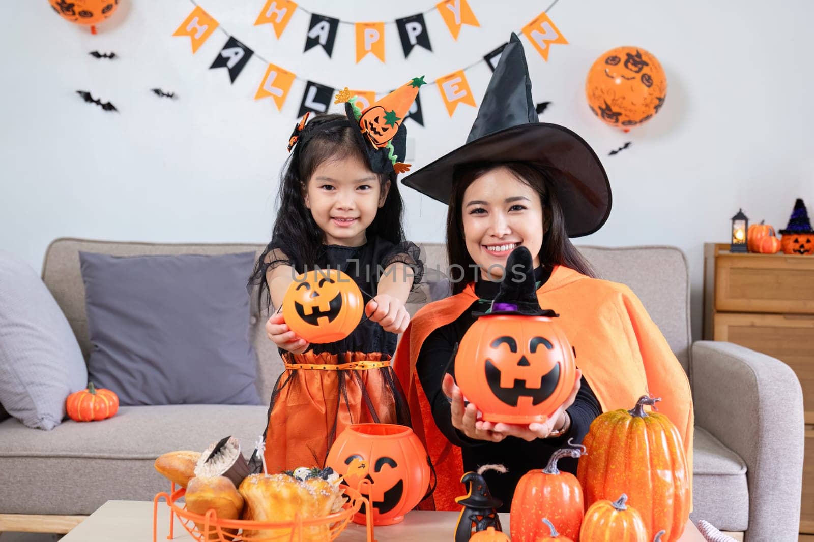 A playful young mother sits on a comfortable sofa in the living room decorated with pumpkins and her cute little daughter tells a spooky Halloween story. Funny mother and girl having fun at home by wichayada