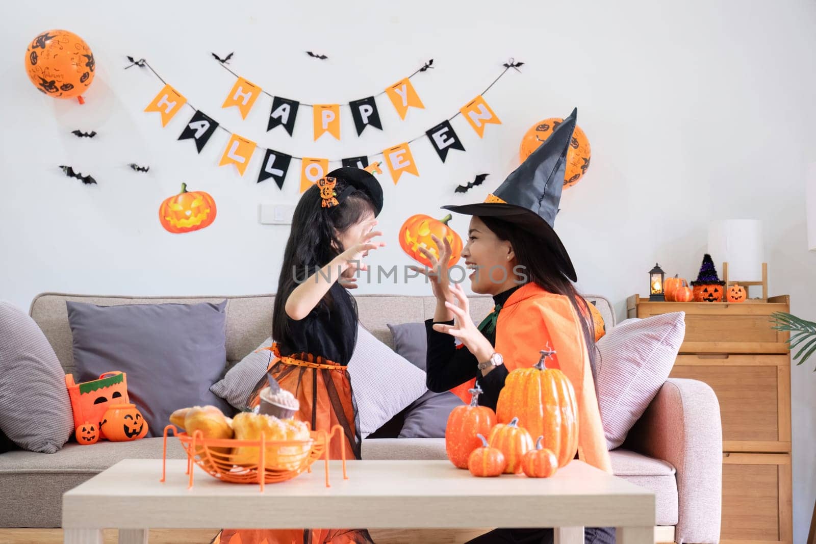 A playful young mother sits on a comfortable sofa in the living room decorated with pumpkins and her cute little daughter tells a spooky Halloween story. Funny mother and girl having fun at home.