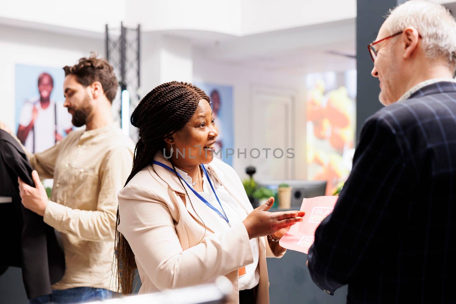 Friendly smiling African American woman shop sales assistant explaining senior man customer Black Friday shopping rules in clothing store. Retail worker holding sales flyer talking with shopper