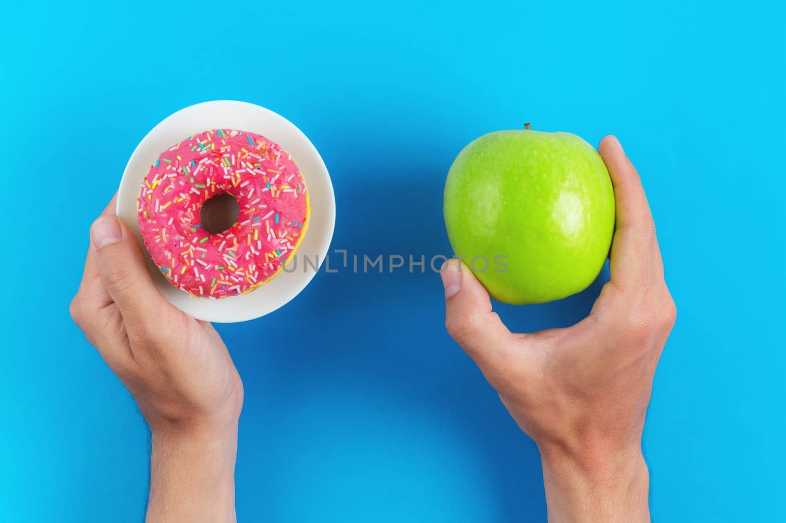 A man chooses between an apple and a donut in his hands. Healthy food concept. Cropped view of male hands holding fresh apple and pink donut offering to make a choice. Healthy food balance concept.