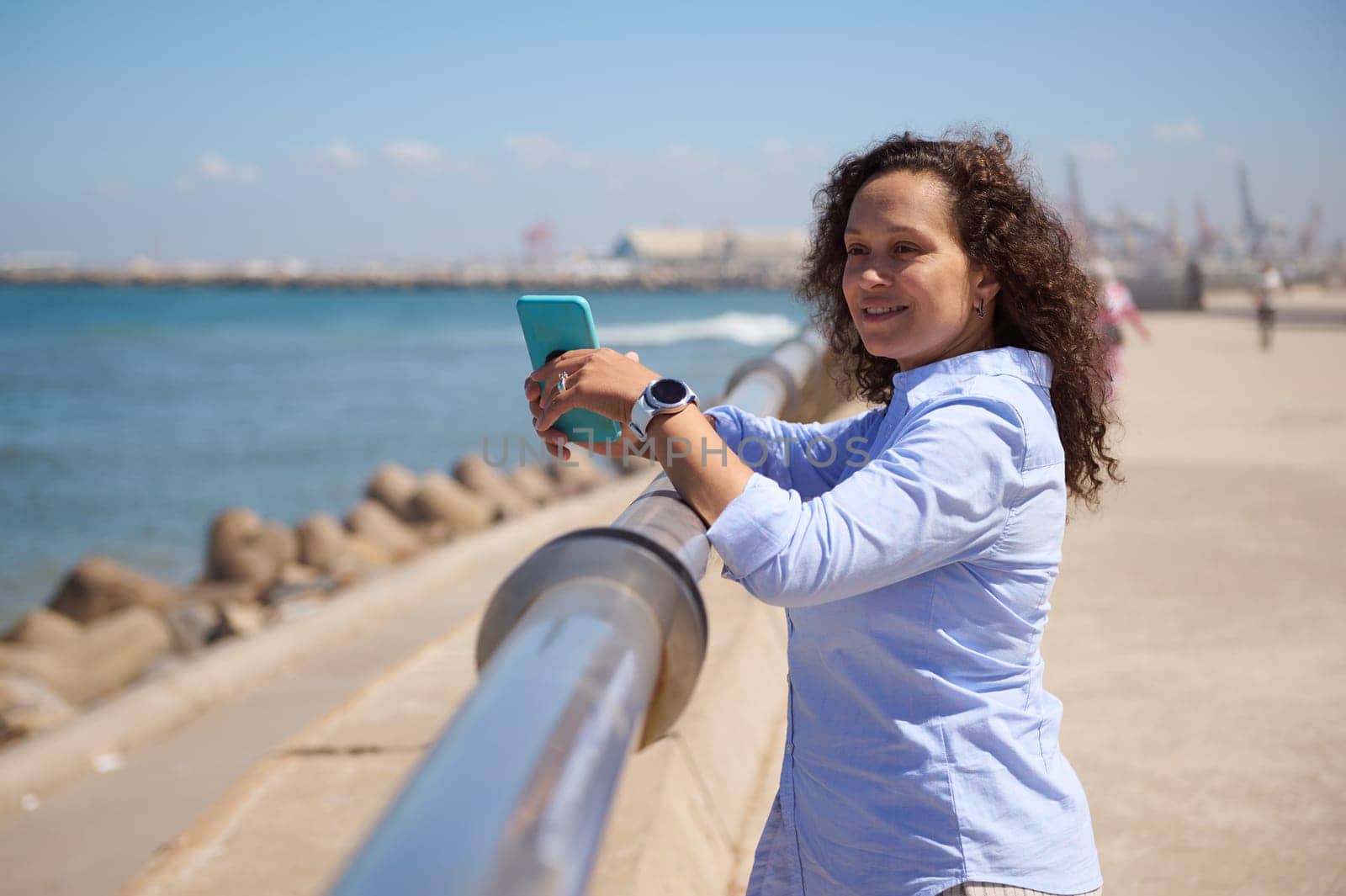 Confident portrait of a smiling pretty woman with mobile phone admiring the beautiful seascape, standing at seacoast on sunny summer day. People. Digital technology. Online communication