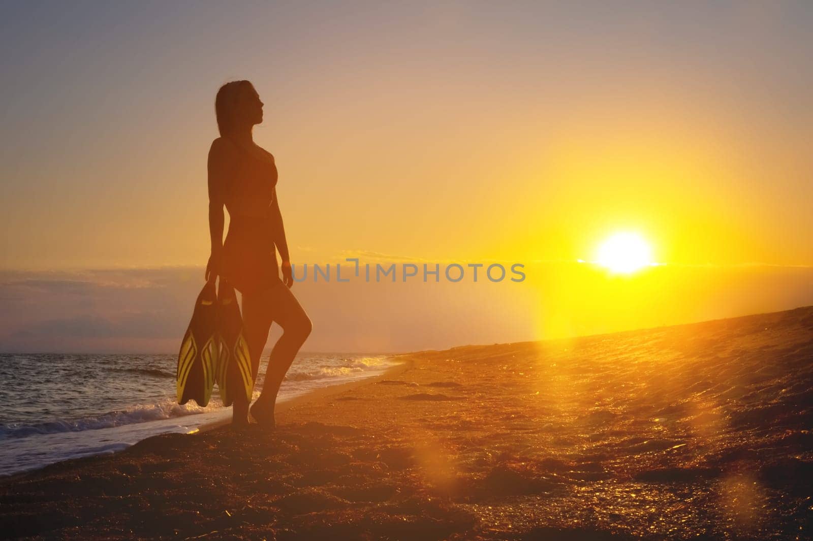 girl diver holds fins in her hands. Summer vacation. Female sexy body silhouette during sunset on vacation after swimming with fins by yanik88