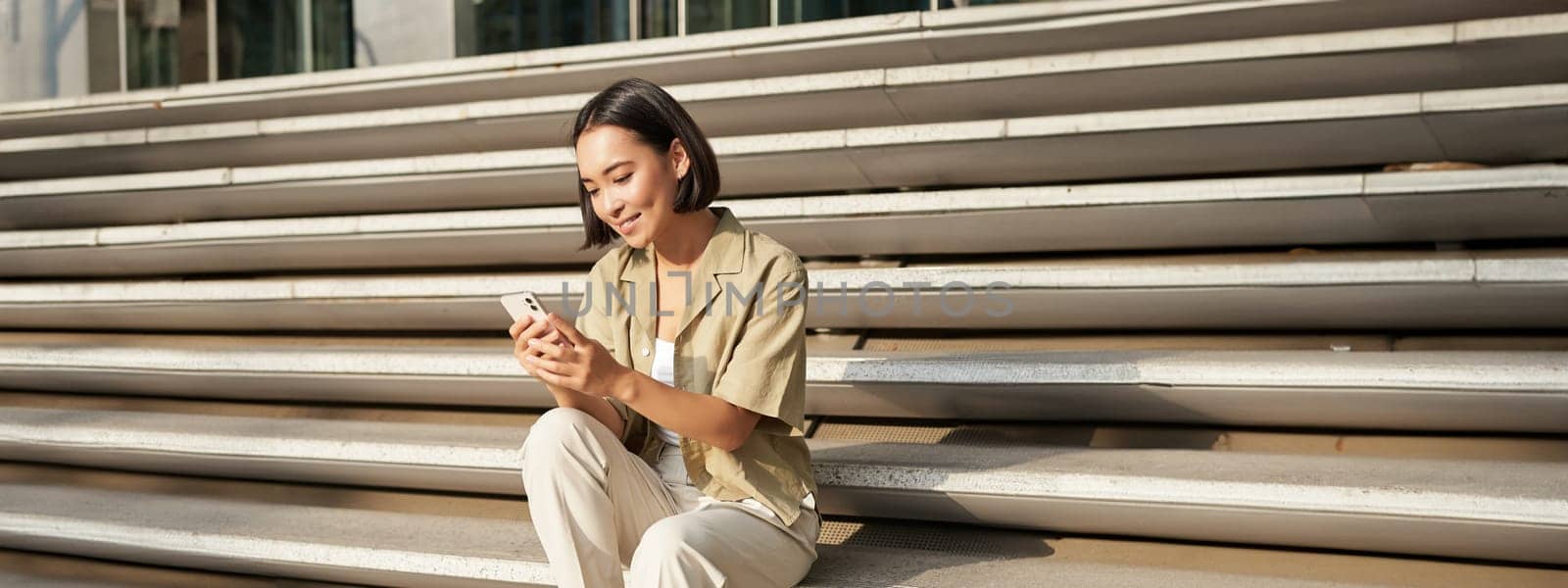 Beautiful asian girl sitting on stairs outside building, using mobile phone, looking at smartphone app and smiling.