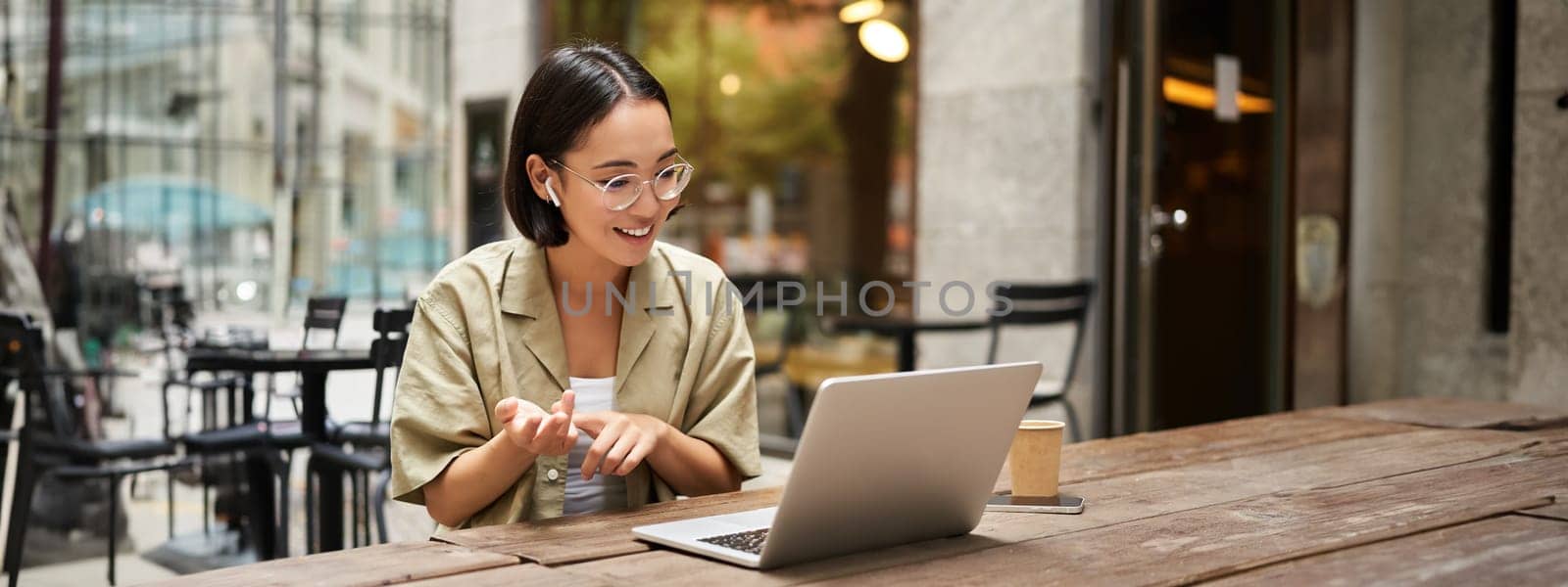 Young woman sitting on online meeting in outdoor cafe, talking to laptop camera, explaining something, drinking coffee. Digital nomad